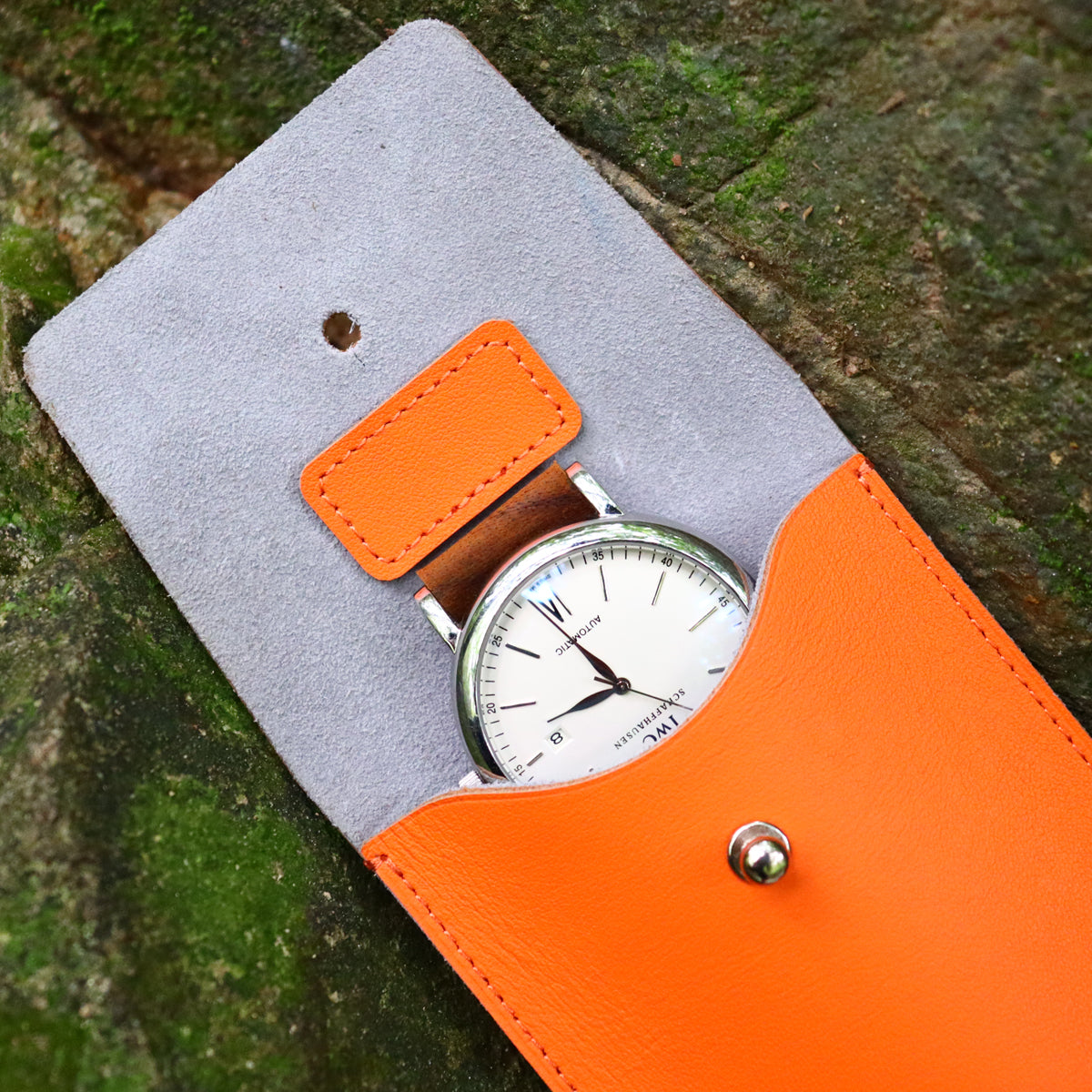 Macro shot of a luxury white dial IWC Schaffhausen automatic mechanical watch being carefully slid into the protective orange leather travel sleeve. The watch strap is neatly threaded through the integrated grey suede insert pad to safeguard the watch case and crystal from friction scratches.