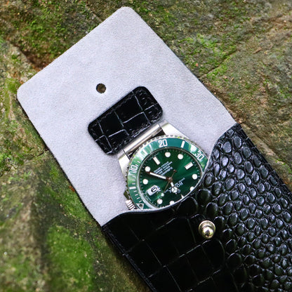 A detailed top-down photograph of a handcrafted, open single-slot watch travel pouch made of high-quality black alligator-embossed leather. The soft, light gray suede-like interior lining and the integrated leather strap holder on the flap are clearly visible. The pouch is empty and resting on a rustic, mossy stone surface, highlighting its textures and construction for luxury watch storage.