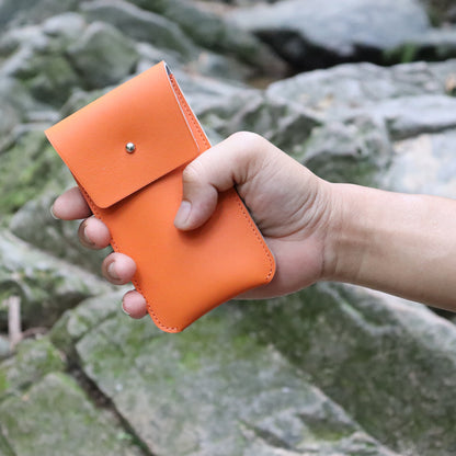 Close-up perspective of a person's hand holding the compact orange leather watch travel pouch in an outdoor environment. The vivid orange color contrasts sharply against a blurred background of natural green mossy rocks, highlighting the pouch’s pocket-sized portability and rugged lifestyle utility