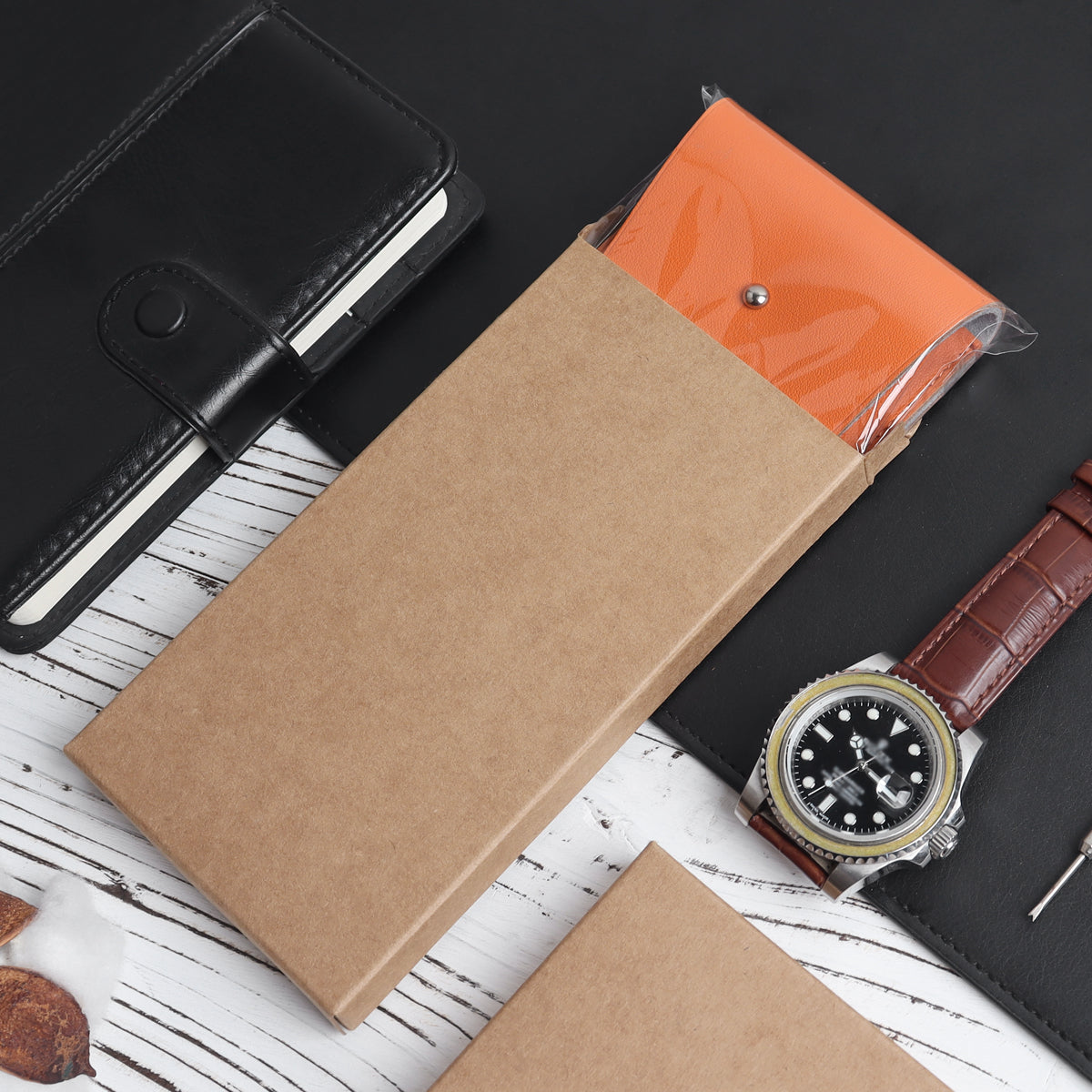 A refined lifestyle flat lay of a gentleman's everyday carry (EDC) setup. The orange leather watch pouch is partially slid out of its minimalist brown kraft paper gift box, styled next to a black leather notebook, a brown alligator-strap dive watch, and a micro screwdriver on a modern white-washed wood grain background.